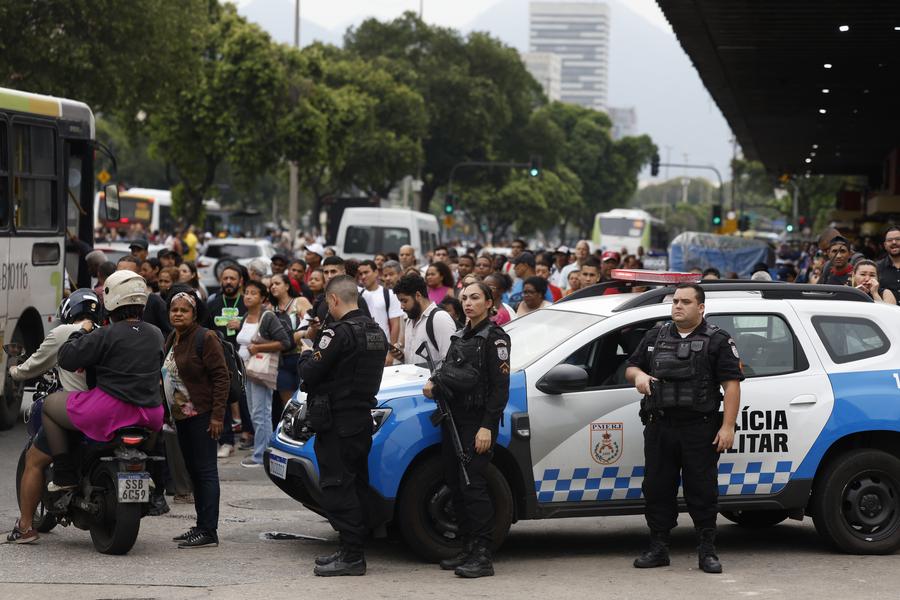 Imagen cedida por Agencia Brasil el 28 de octubre de 2025 de oficiales de policía montando guardia mientras personas esperan en una parada de autobús durante una megaoperación de las fuerzas de seguridad contra un grupo criminal, en Río de Janeiro, Brasil. (Xinhua/Fernando Frazao/Agencia Brasil)